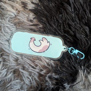May include: A rectangular acrylic keychain with rounded edges featuring a light blue background and a central graphic of a stylized, curved pink and white creature. The keychain includes a light blue plastic lobster clasp attachment.