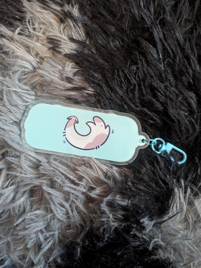 May include: A rectangular acrylic keychain with rounded edges featuring a light blue background and a central graphic of a stylized, curved pink and white creature. The keychain includes a light blue plastic lobster clasp attachment.