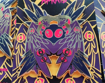 May include: A purple and gold enamel pin featuring a stylized mothman design with pink accents. The pin has the text "BE MOTH AFRAID" and "Biblically Accurate Mothman". The pin is on a black and purple patterned backing card.
