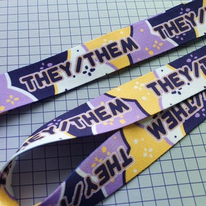 May include: Two lanyards with a repeating pattern of the words "THEY/THEM" in a dark blue, stylized font. The background features a mix of white, yellow, and purple geometric shapes. The lanyards are on a grid background.