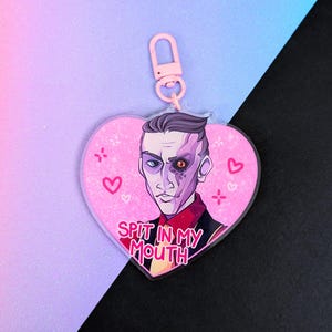 May include: A heart-shaped acrylic keychain featuring a stylized illustration of a person with a split-tone face, one side purple and one side skin-toned, wearing a red shirt and yellow tie, with the text SPIT IN MY MOUTH and pink heart accents.
