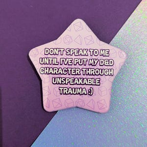 Op de afbeelding: Een stervormige pin met een lichtpaarse gradiënt en de tekst "DON'T SPEAK TO ME UNTIL I'VE PUT MY D&D CHARACTER THROUGH UNSPEAKABLE TRAUMA :) ". De pin heeft een patroon van dobbelstenen en geometrische vormen.