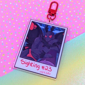 Pode incluir: Um porta-chaves de acrílico transparente com uma ilustração de desenho animado de uma criatura semelhante a uma traça com olhos vermelhos e asas pretas. O porta-chaves tem um fecho vermelho e o texto "Sighting #23 10/12/20" em vermelho. O fundo é rosa e azul.