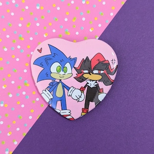 May include: A heart-shaped button featuring cartoon characters Sonic and Shadow holding hands. Sonic is blue with green eyes, and Shadow is black and red. The background is pink with a small red heart.