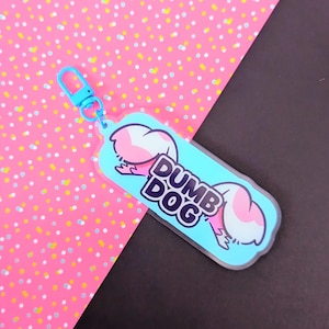 May include: A light blue acrylic keychain featuring a cartoon illustration of a pink and white dog tail and the text DUMB DOG in bold black letters. The keychain includes a matching light blue plastic lobster clasp for attachment to items.
