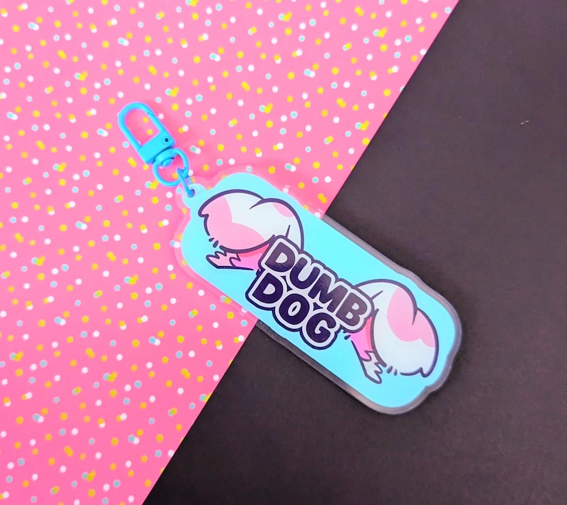May include: A light blue acrylic keychain featuring a cartoon illustration of a pink and white dog tail and the text DUMB DOG in bold black letters. The keychain includes a matching light blue plastic lobster clasp for attachment to items.