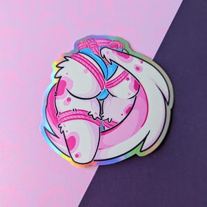 Può includere: Un adesivo olografico con un'illustrazione a cartoni animati di una creatura con pelle bianca e rosa, legata con una corda rosa. La creatura ha grandi tratti arrotondati. Lo sfondo è rosa e viola.