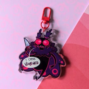 May include: A clear acrylic keychain featuring a cartoon moth with large pink eyes, holding a knife. The moth is purple and black with pink accents. Text on the keychain reads "I CRAVE VIOLENCE". The keychain has a red clasp.