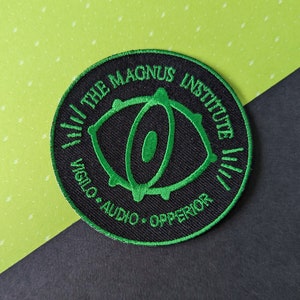 May include: Black and green embroidered patch with the text "The Magnus Institute" and "Visilo Audio - Operator" in a circle around a stylized eye graphic.