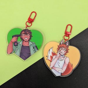 May include: Two heart-shaped keychains with red clasps. One features a cartoon man holding a drill to his head, set against a green background. The other depicts a fox-eared character with a white top and orange background.