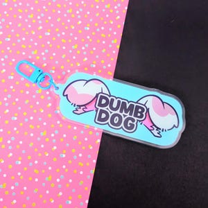 May include: A rectangular acrylic keychain with rounded corners featuring a light blue background and the text DUMB DOG in bold black letters. The design includes two stylized pink and white animal ears on either side and a light blue lobster clasp.