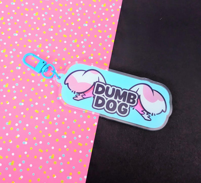 May include: A rectangular acrylic keychain with rounded corners featuring a light blue background and the text DUMB DOG in bold black letters. The design includes two stylized pink and white animal ears on either side and a light blue lobster clasp.