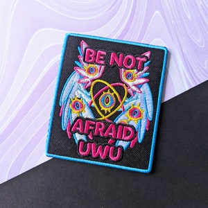 Peut inclure: Patch brodé noir avec un motif rose et bleu représentant deux yeux ailés et le texte "BE NOT AFRAID UWU".