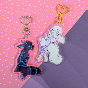 May include: Two acrylic keychains featuring anthropomorphic characters with heart-shaped clasps. One keychain displays a black character with blue accents and blue rope bindings, while the other shows a white character with purple rope bindings.