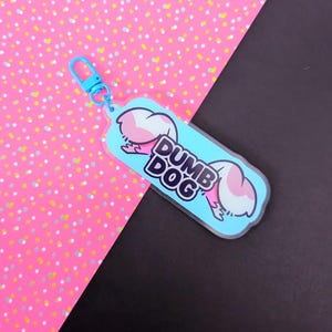 May include: A light blue acrylic keychain featuring a cartoon illustration of a dog with pink ears and the text DUMB DOG in bold black letters. The keychain includes a matching light blue plastic lobster clasp attached to the top of the charm.