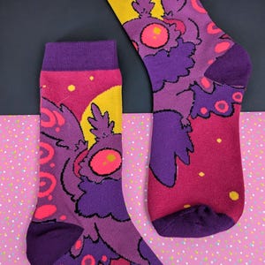 Peut inclure: Une paire de chaussettes montantes arborant un motif graphique coloré représentant une créature ressemblant à un papillon de nuit violet sur un fond rose et jaune. Les chaussettes présentent un revers, un talon et une pointe violet foncé.
