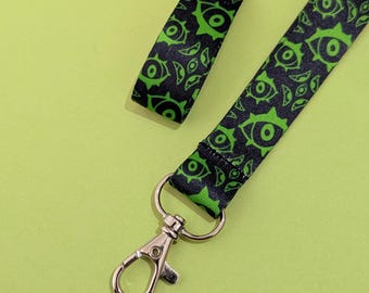 May include: A black and green lanyard with a repeating eye pattern. The lanyard has a silver-colored metal clasp and a swivel hook. The background is a solid light green color.
