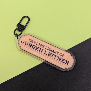 May include: A rectangular keychain with the text "FROM THE LIBRARY OF JURGEN LEITNER" in a vintage style. The keychain has a black clasp and a clear body with a peach-colored label.
