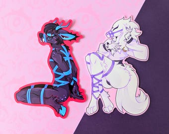 May include: Two stickers featuring stylized anthropomorphic creatures. One is dark purple with blue accents and restraints, the other is white with purple restraints. Both are on a pink and purple background.