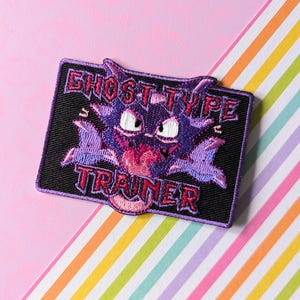 Könnte beinhalten: Gestickter Aufnäher mit einem lila Cartoon-Monster mit dem Text "GHOST TYPE TRAINER" in Pink und Lila. Der Aufnäher befindet sich auf einem schwarzen Hintergrund mit einem lila Rand. Der Hintergrund ist ein rosa und gestreiftes Muster.