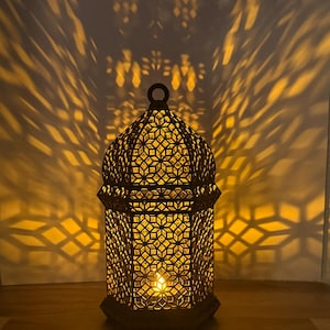 May include: A dark-colored metal lantern with an intricate geometric cutout pattern. The lantern features a dome-shaped top with a small ring handle and emits a warm, glowing light from within, casting complex star-shaped shadows on the wall behind.