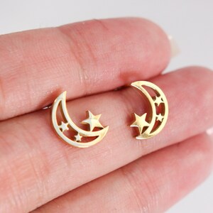 May include: A pair of gold-colored stud earrings. Each earring features a crescent moon shape with cut-out stars. The earrings are small and delicate, with a polished finish. The earrings are designed to be worn in pierced ears.