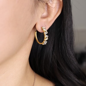Peut inclure: Boucles d'oreilles créoles dorées avec une rangée de pierres claires en forme de baguette.