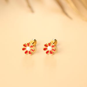 May include: A pair of gold-colored stud earrings. Each earring features a red and white daisy design. The petals are red with a gold outline, and the center is white. The earrings are set against a light beige background.