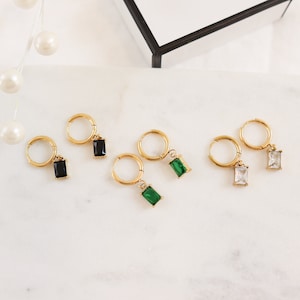 May include: Three pairs of gold hoop earrings with dangling charms. The charms are rectangular and feature green, black, and clear gemstones.