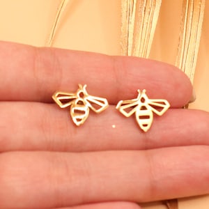 May include: A pair of gold-colored bee-shaped stud earrings. The earrings feature a geometric design with wings and body details. The earrings are small and delicate, suitable for everyday wear.