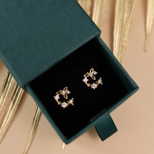 May include: A pair of gold earrings with a delicate floral design and clear cubic zirconia stones. The earrings are displayed in a green velvet jewelry box.