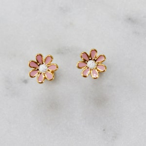 Könnte beinhalten: Ein Paar Stecker-Ohrringe mit Daisy-Motiv. Jeder Ohrring hat eine rosa Emailleblume mit goldfarbenem Rand und einem weißen Zentrum. Die Ohrringe sind vor einem weißen Marmorhintergrund platziert.