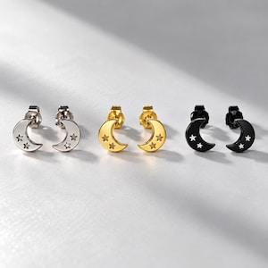 May include: Three pairs of stud earrings shaped like crescent moons, each featuring two small star cutouts. The set includes one pair in silver tone, one pair in gold tone, and one pair in black tone, all with standard post backings.