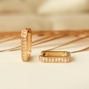 May include: Two gold-colored hoop earrings with rectangular shapes. Each earring is adorned with a row of clear, rectangular-cut gemstones. The earrings are displayed on a light-colored surface with a blurred background.