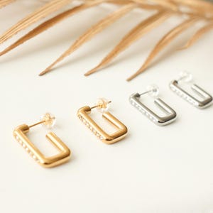 May include: Three pairs of earrings with a square shape and a row of clear rhinestones. The earrings are in gold, silver, and rose gold.