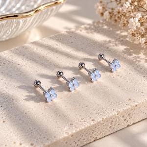 May include: Four silver-toned barbell earrings, each featuring a small flower-shaped charm composed of four translucent, pale blue, rounded stones. The earrings have a straight post with a small spherical backing, designed for piercing.