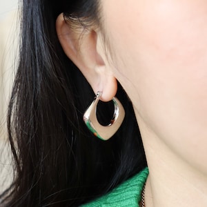 Puede incluir: Pendientes de aro cuadrados de plata con un borde ligeramente redondeado. Los pendientes se llevan en la oreja de una persona.