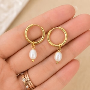 May include: A pair of gold-toned hoop earrings, each featuring a single small, oval-shaped white pearl drop suspended from the bottom. The earrings have a polished metallic finish and a simple hinged closure mechanism for secure wear on the earlobe.