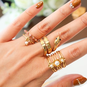 Op de afbeelding: Een hand met een set gouden ringen, waaronder een slangenring met zwarte ogen, een ring met een parel en een ring met roze kralen.