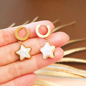 May include: A pair of gold hoop earrings with a white star charm. The stars have an iridescent finish.