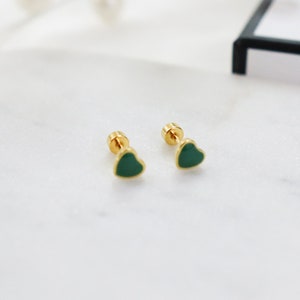 May include: A pair of gold-colored stud earrings, each featuring a heart-shaped, dark green enamel design. The earrings are displayed on a white marble surface, showcasing their polished finish and simple elegance. The earrings are small and delicate.
