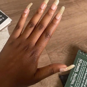 Puede incluir: Una mano con uñas largas, de color marrón claro y forma ovalada. Las uñas son largas y se extienden más allá de las yemas de los dedos.