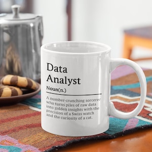 May include: A white ceramic mug featuring black printed text that reads Data Analyst Noun(n.) followed by a definition describing a number-crunching sorcerer who turns raw data into golden insights with the precision of a Swiss watch and a cat.