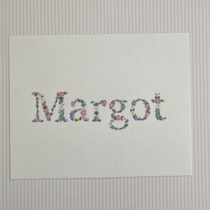 May include: A white card with the name "Margot" spelled out in floral embroidery. The flowers are pink, blue, and yellow.