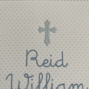 May include: White fabric with a blue embroidered cross, the name Reid William, and the word "Baby" in a cursive font.
