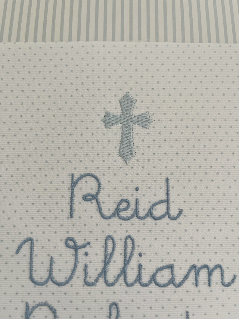 May include: White fabric with a blue embroidered cross, the name Reid William, and the word "Baby" in a cursive font.