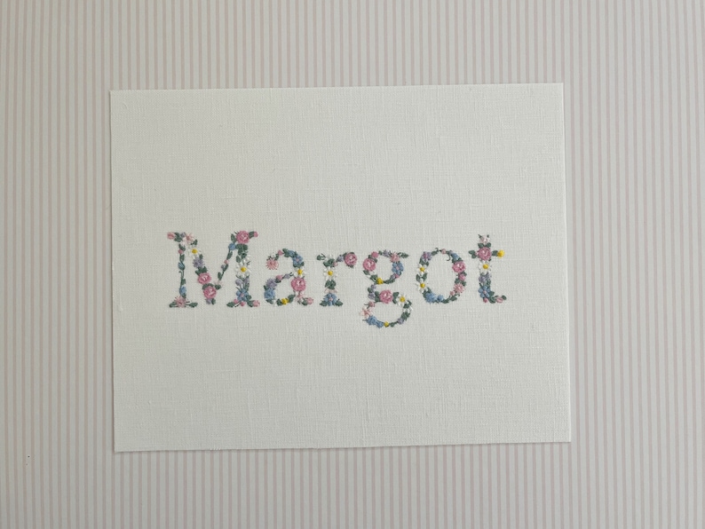 May include: A white card with the name "Margot" spelled out in floral embroidery. The flowers are pink, blue, and yellow.