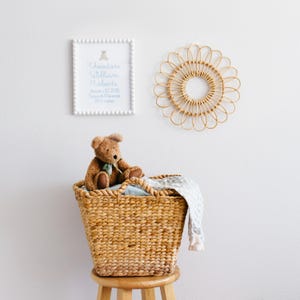 May include: A nursery setting with a woven basket on a wooden stool, containing a teddy bear and a blanket. A framed print with baby's name and birth details is on the wall, alongside a rattan wall decoration. The print reads: Theodore William Roberts, January 27 2025, 3.4 kg, 51 cm.