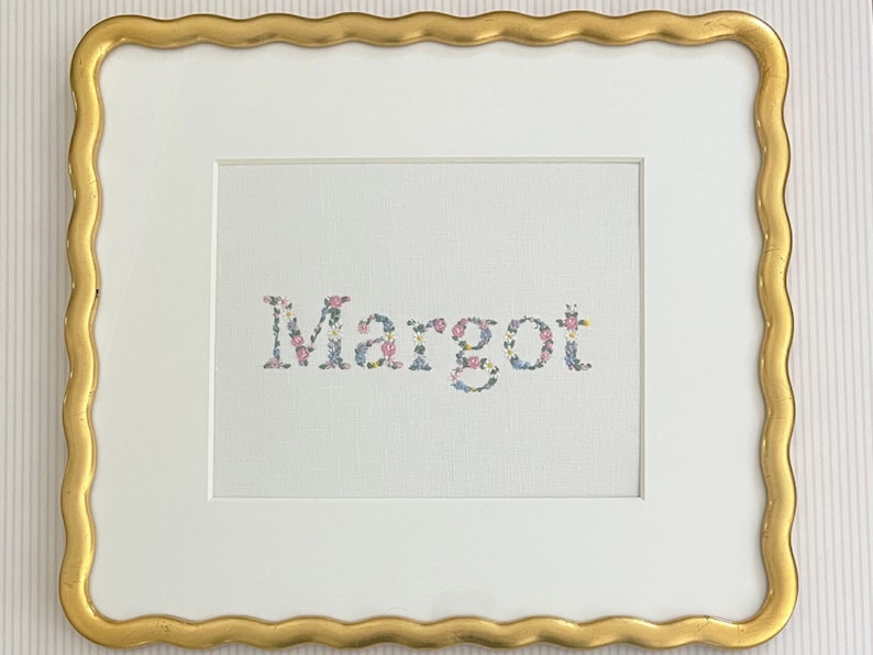 May include: A white frame with a gold scalloped edge surrounds a white background with the name "Margot" spelled out in colorful flowers.