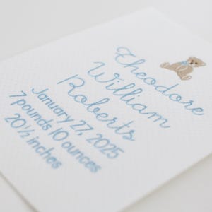 May include: A white card with light blue embroidered text and a small teddy bear illustration. The text reads "Theodore William Roberts, January 27, 2025, 3.18 kg, 52.07 cm". The card has a subtle dotted pattern.